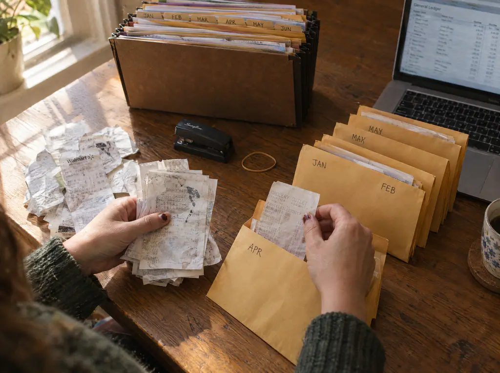 Hands sorting paper receipts into monthly manila envelopes next to an accordion file